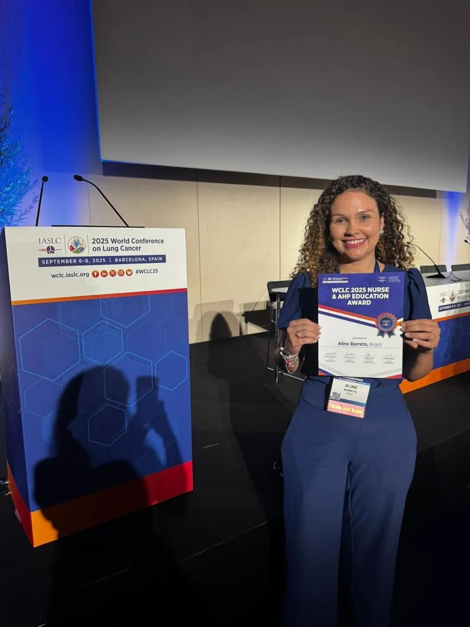 Aline Barcellos segurando o certificado do prêmio ao lado do púlpito da 2025 World Conference on Lung Cancer